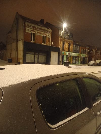 A nighttime scene shows a parked car covered in snow on the right. To the left, a streetlight illuminates a brick building with windows, awnings, and architectural details. The streetscape includes businesses with visible signs, indicating a commercial area. The sky is dark, suggesting a late hour or heavy cloud cover. The overall image is slightly rotated.