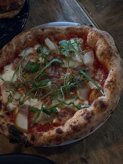 A close-up, overhead shot depicts a pizza on a white plate set on a wooden table. The pizza has a raised crust with browned spots, and is topped with tomato sauce, cheese, thinly sliced pears, cured meat, and a garnish of fresh green leaves (likely arugula). Part of a blue and black plate with food is visible in the lower left corner of the frame and part of a black plate is visible in the lower right. The location is Tlaxcala City, Mexico.