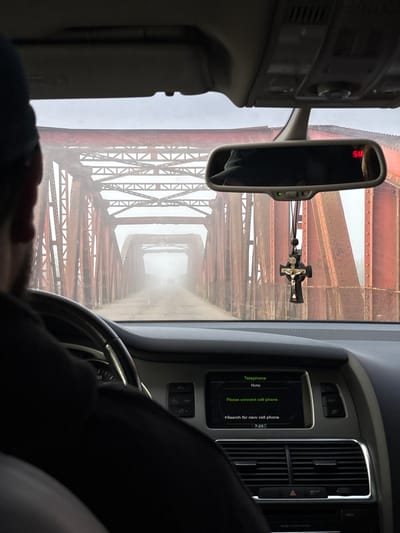 The image shows the interior of a car driving across a bridge. The bridge is made of red, rusted metal trusses and is covered in fog. The road surface is visible and appears to be dirt or gravel. The dashboard of the car is visible, with a display screen showing a "Telephone" menu and the time "7:23". A Christian crucifix is hanging from the rearview mirror. The scene suggests a journey through a rural or industrial area, with the fog adding an element of mystery or isolation. The location is identified as Dibrova, Ukraine.