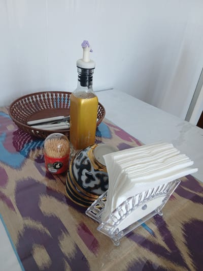 An indoor scene at a restaurant in Gulistan, Uzbekistan. A table is covered with a patterned cloth. On the table are several items, including a brown woven basket containing cutlery wrapped in white paper, a clear bottle of golden liquid with a purple dispenser cap, a cylindrical dispenser filled with toothpicks, a decorative bowl with black, gold, and white intricate patterns, and a clear plastic napkin holder filled with white paper napkins. The background consists of a white wall.