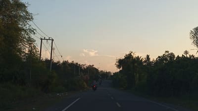 This image depicts a road scene at dusk, likely in Tanjongkeumala, Indonesia. A paved road curves slightly into the distance, flanked by lush green vegetation and trees on either side. Utility poles with power lines are visible on the left side of the road. In the foreground, a motorcycle with a rider is traveling away from the camera. A few other motorcycles are visible further down the road. The sky is a soft gradient of blue and orange, indicative of sunset, with a few scattered clouds. The overall impression is of a rural or semi-rural road at the end of the day.