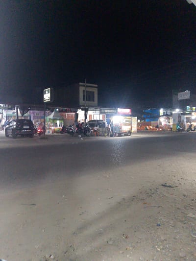 This media file captures a street scene at night in Gampongpaya, Indonesia. The image is dimly lit, suggesting it was taken after sunset. The foreground shows a paved road with scattered debris, leading towards a row of small shops and food stalls illuminated by artificial lights.
Several vehicles are visible, including a black car parked on the left side of the road, a scooter in the middle, and motorcycles further down the street. There are also a few people present, some near the vehicles and others gathered around the food stalls. The overall atmosphere appears to be that of a typical evening gathering place, with activity centered around the commercial establishments.
One of the stalls displays the text "WM BOUTIQUE" prominently on a sign, indicating a retail business. Other signs and text are present on the stalls, but they are difficult to read due to the low light and image quality. The weather appears to be clear, as there are no indications of rain or adverse conditions. The scene evokes a sense of everyday life and commerce in a semi-urban or rural Indonesian setting at night.