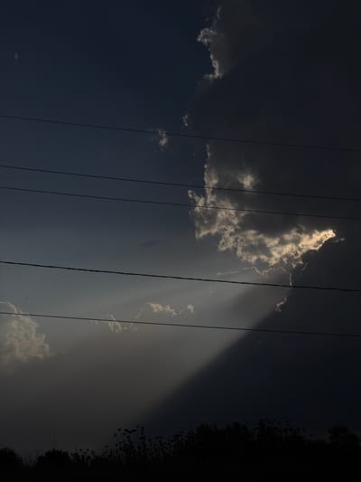 The image shows a sky filled with dramatic clouds, illuminated by shafts of sunlight. In the foreground, a silhouetted line of trees and tall grass runs along the bottom of the frame. Four parallel power lines stretch horizontally across the sky. The overall impression is one of a dramatic and moody sky, possibly just before or after a storm. The location is identified as Marneuli, Georgia.