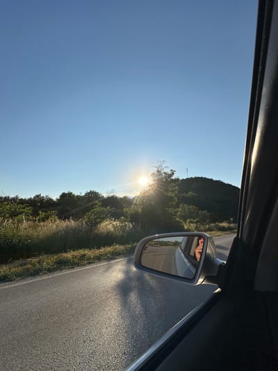 The image depicts a view from inside a car looking out the passenger side window, with the car in motion on a paved road. The scene is set during the day, likely late afternoon or early evening due to the low angle of the sun and the warm, golden light it casts. The sun is bright and appears to be setting or rising, creating a lens flare and illuminating the surrounding landscape.
The roadside is lined with lush green vegetation, including trees and tall grasses, suggesting a rural or semi-rural location, consistent with the provided location context of Prisovo, Bulgaria. A hillside covered in trees is visible in the distance. A rearview mirror is mounted on the passenger side door, reflecting a partial view of the driver's face, specifically their eyes and forehead, and what appears to be their hands resting on the steering wheel. The overall mood is serene, possibly conveying a sense of travel or contemplation. The sky is a clear, bright blue, indicating fair weather. There is no visible text in the image.