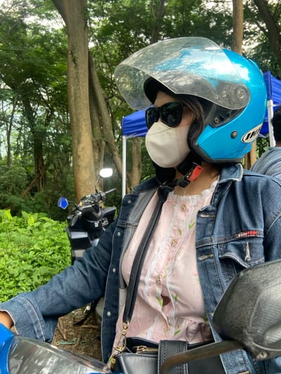A person wearing a blue helmet with a clear visor, sunglasses, and a white face mask sits on a motorcycle. The individual is also wearing a denim jacket over a light pink floral-patterned shirt with buttons and lace trim. A black purse hangs to the side. The motorcycle is parked in a grassy area with trees in the background.