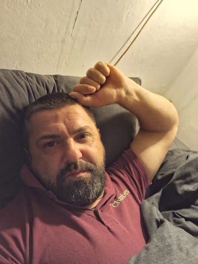 A man with a beard lies on a pillow in bed. He is wearing a burgundy polo shirt with the name "Chambers" embroidered on it. He is covered by a gray blanket. His right arm is raised and his hand is clenched into a fist. The location is not possible to determine based on the image.