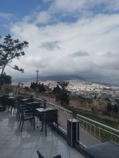 The image captures a cityscape of Tétouan, Morocco, under a sky with partial cloud cover. A mountain is visible in the background, partially obscured by clouds. In the foreground, there is an outdoor terrace with black chairs and tables arranged neatly on a tiled surface. A decorative railing separates the terrace from a road and lower-lying buildings. A communication tower stands amidst the urban sprawl. The scene appears to be captured from an elevated vantage point, possibly a restaurant or cafe.