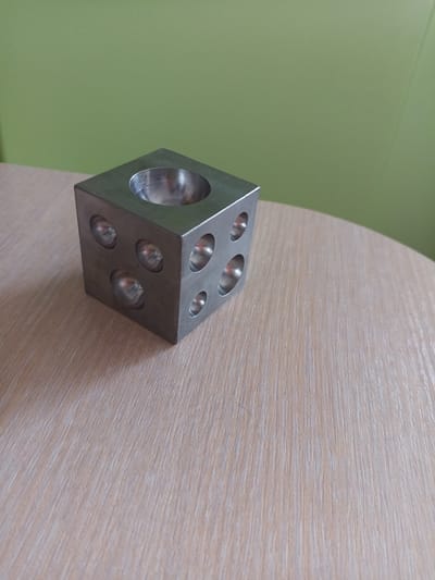 A metal cube with indentations on its faces sits on a wooden table. The cube has a circular indentation on its top face and multiple circular indentations on its visible side faces. The background is a plain green wall. The image is a still life photograph with no people or actions depicted. There is no information in the image to suggest a specific location in Vilyuysk, Russia.
