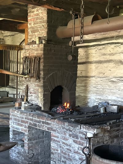 This is a photograph of a blacksmith's forge, likely in a historical setting in South Dundas, Canada. The scene is dimly lit, suggesting either indoor conditions or evening time. The focal point is the forge itself, a brick structure with a glowing fire at its heart, indicated by visible embers and flames. Tools of the trade, such as tongs and hammers, are neatly arranged on a brick wall to the left of the forge, suggesting active use. A large, tan-colored pipe extends from the upper left of the image, possibly part of a ventilation or heating system. Chains and metal fixtures are also visible, adding to the industrial feel of the space. The walls are rough-hewn brick and possibly wood, contributing to the rustic and historical ambiance. The overall impression is one of a functional workspace, steeped in the tradition of blacksmithing. There is no visible text.