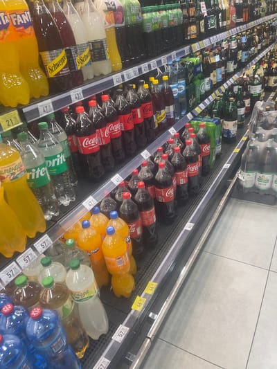 This image depicts a grocery store aisle in Gozhuly, Ukraine, filled with a diverse selection of bottled beverages. The shelves are densely packed with a variety of soft drinks, including Coca-Cola, Fanta, and Sprite, alongside juices and other bottled beverages. The lighting suggests an indoor retail environment. There are no people visible in the image, only the merchandise on display. The overall scene is one of commercial retail stock presentation.