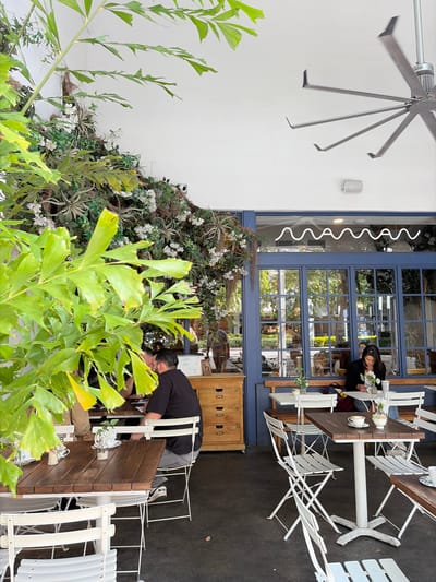 A scene in Coral Gables, USA, features a restaurant patio partially obscured by lush green foliage. A white wall is adorned with climbing plants and white flowers. A large, modern metal fan is mounted on the wall. Two people are seated at tables. One woman is sitting at a table indoors. The second person is outdoors. They appear to be interacting with a third person who seems to be rearranging chairs. White bistro-style chairs are positioned around dark wood tables. Decorative lighting, a blue-framed window, and wooden cabinetry are also present.