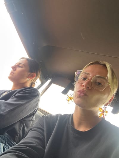 The image captures two women inside what appears to be a vehicle, likely a safari or transport jeep, with a dark, possibly fabric or vinyl, overhead roof structure visible. The shot is taken from a low angle, looking upwards.

In the foreground, a blonde woman with eyeglasses is looking directly at the camera, making a playful pouting expression. She is wearing a dark top and large, amber-colored, flower-shaped earrings. Her face and part of her torso are visible in the upper right section of the frame. Below and to her left, a second woman with dark hair is seen in profile, looking downwards. She also wears a dark top and a small, metallic earring.

A significant portion of the lower and central part of the image is filled with bright white light, suggesting a clear sky or intense daylight from an open side or window of the vehicle. The overall lighting indicates it is daytime. No discernible text is visible. The scene's context, Situlpawwa, Sri Lanka, further supports the interpretation of a safari or tour vehicle in a natural or historical setting.