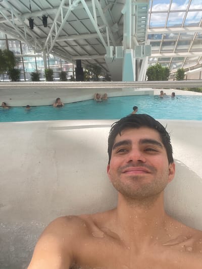 The image depicts a man in a swimming pool, possibly at Caldea, a thermal spa in Les Escaldes, Andorra. The man's face is close to the camera, with visible water droplets on his skin. The background shows other people in the pool, and the spa's distinctive glass and steel structure, typical of Caldea's architecture. The image is taken from an unusual angle, rotated so the pool appears vertical.