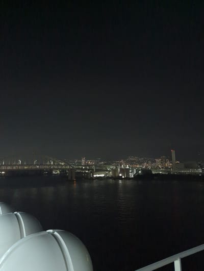 The image captures a nighttime view of the Kobe waterfront, likely from a ship or ferry. In the foreground, a portion of a white, curved object, possibly a life raft or part of the vessel's structure, is visible. The middle ground features the dark, rippling water of the harbor, reflecting the distant city lights. Across the water, the illuminated Kobe cityscape stretches along the coast. A prominent bridge, appearing to be the Kobe Ohashi Bridge or a similar structure, is visible, with its lights forming a long, luminous line. Beyond the bridge, numerous buildings and lights of the city create a sprawling urban panorama against the dark sky. A tall, white tower with a red light at its apex stands out among the city buildings, possibly a landmark like a lighthouse or communication tower. Industrial structures and port facilities are also discernible along the waterfront, suggesting a bustling port area. The overall scene conveys a sense of a vibrant city at night, viewed from the calm waters of its harbor.