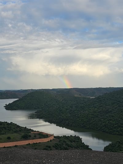 The image depicts a panoramic view of a reservoir or large river winding through verdant, undulating hills in Oumazza, Morocco, under an overcast sky. A prominent rainbow is visible in the upper center of the frame, arcing across the sky against a backdrop of varied cloud cover, ranging from lighter white to darker grey masses. The foreground consists of a dark, gravelly slope, suggesting elevated terrain from which the photograph was taken. In the midground, a low, light orange-brown wall or barrier with a slight curve runs along a section of land bordering the water, separating it from sparse green vegetation. The water itself is dark, reflecting the sky and surrounding hills, and appears calm. The numerous hills surrounding the water body are densely covered with dark green vegetation. No people, specific subjects beyond the landscape elements, actions, or interactions are visible.