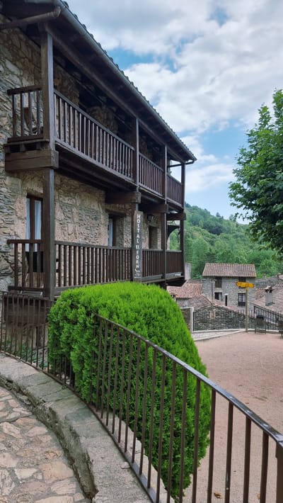 The image shows a rustic stone building with wooden balconies and railings, likely an inn or guesthouse, situated in a hilly, green landscape. A prominent sign on the building reads "HOSTAL EL FORN". In the foreground, a neatly trimmed green bush is visible behind a metal fence. Other stone buildings with tiled roofs, typical of a Spanish village, are visible in the background, along with a tree-covered hillside. The sky is partly cloudy. The setting suggests a rural or semi-rural environment within Camprodon, Spain. There are no people visible in the image.