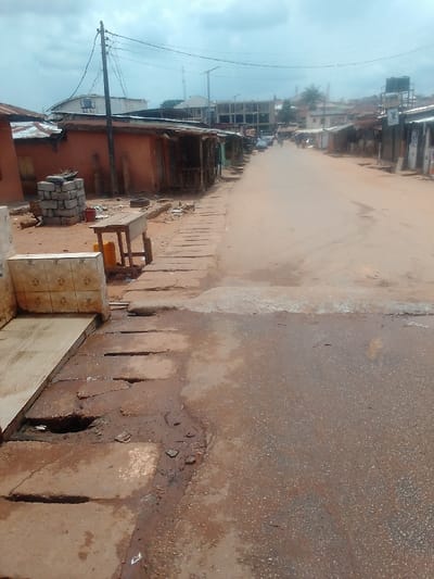 The image shows a narrow, dirt road in Auchi, Nigeria, located at coordinates 7.063 latitude and 6.261 longitude. The road is lined with small, one-story buildings on both sides. The buildings appear to be made of concrete or brick and have corrugated metal roofs. There are several power lines running along the street. In the foreground, there is a concrete drainage ditch running along the side of the road. There are a few people walking along the street in the distance, but none are visible in the foreground. The scene appears to be taken on a cloudy day. The road is dusty and there is debris scattered on the ground. The image is likely taken from a pedestrian's perspective, looking down the street.