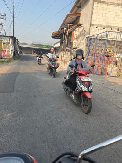 In Karangtanjung, Indonesia, a street scene unfolds with multiple individuals riding motorcycles. The foreground features a red and black Honda motorcycle, license plate DK 3588 LP, with a person wearing a denim jacket, pink headscarf, and white face mask at the handlebars. Behind them, another motorcycle is partially visible. Further down the road, a second motorcycle, a red Honda, carries a person in a black head covering and pink pants. In the distance, a third motorcycle is seen. The environment includes buildings, including one with a sign that reads "LOSS GYM MULYO," power lines, and a clear sky. The road surface appears to be asphalt. Construction materials, including stacked bricks and scaffolding, are visible on the right side of the road. The lighting suggests it is daytime.