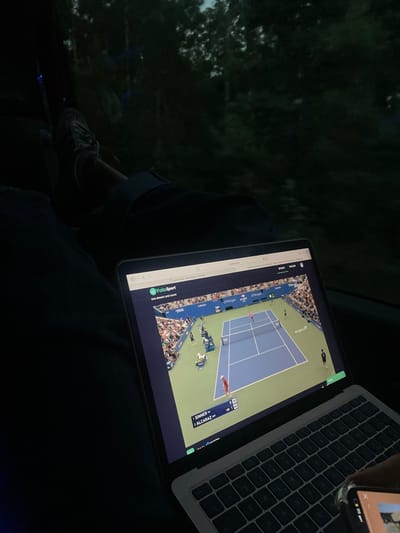 The scene is set in an interior location, consistent with a vehicle, during low light conditions in Albergaria-a-Velha, Portugal. An individual's legs are visible, clad in dark trousers and light-toned athletic shoes, with one foot elevated. A silver Apple MacBook laptop occupies the foreground, its screen illuminated and displaying a live stream of a tennis match on the YoloSport website. The match features a blue tennis court, two players, and spectators in the stands, with a digital scoreboard showing "1 SINNER 0" and "2 ALCARAZ 0," accompanied by a "49" minute timer. A smartphone is partially visible in the lower right, held by a hand, with its screen displaying an image. The background, viewed through a window, reveals dark, indistinct foliage, indicating an outdoor environment.