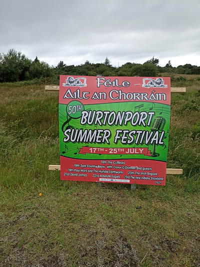 A large, red and green poster advertising the "BURTONPORT SUMMER FESTIVAL" is mounted on wooden planks outdoors. The poster features a vintage microphone graphic, musical notes, and text in English and Irish. The sky is overcast, and the poster is situated in a grassy area with trees in the background. The bottom of the poster lists various events and performers, including "19th 3pm Country Show with Conor O'Donnell and guests," "19th Paul Ward and The Humble Earthworm," "20th The Irish Brigade," "21st David James," and "22nd Absolute Eagles," and "23rd The new Indians Showband." The festival is scheduled to take place from the 17th to the 25th of July.