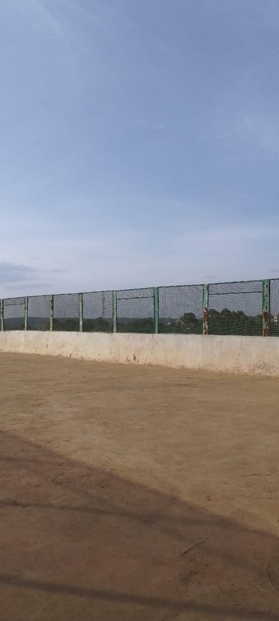 The image captures an outdoor scene dominated by a vast sky and an expanse of dry ground, separated by a fence and a concrete wall.

The upper portion of the image features a clear, light blue sky with minimal, very faint wispy cloud formations. The lighting suggests it is daytime, likely a sunny period.

Horizontally across the middle of the frame, a long fence structure divides the sky from the foreground. This fence is composed of a dark, fine-mesh chain-link material, supported by regularly spaced vertical metal posts. The posts appear to be painted a faded or weathered green, with some areas showing signs of rust or chipped paint, revealing a reddish-brown color underneath, particularly noticeable on the right side. The fence itself stands on top of a sturdy, light-colored concrete wall or barrier, which also runs horizontally across the scene. This concrete wall shows some discoloration and rough textures, indicating age or exposure to elements. Behind the fence, a strip of darker green is visible, likely representing distant vegetation, trees, or hills, which are somewhat obscured by the fence mesh.

The lower half of the image is filled with an unpaved, reddish-brown ground surface, appearing dry and somewhat dusty, resembling dirt or sand. There are subtle variations in the ground's texture and color, with some faint indentations or patterns, possibly from foot traffic or vehicle marks. Long, dark shadows stretch diagonally across the ground from the left side towards the right, indicating the sun is positioned to the right of the frame, suggesting either morning or late afternoon.

Overall, the scene depicts a static, open-air environment, possibly a perimeter of a field, an undeveloped plot of land, or an area awaiting construction, under a bright, clear sky. No human activity or specific event is taking place, and no text is visible in the image.