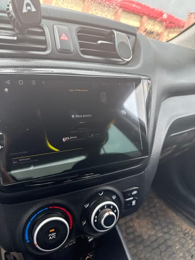 The image shows the interior of a car. The focus is on the center console, which includes a touchscreen display showing a music application, likely "Yandex Music," as indicated by the text "Яндекс Музыка" (Yandex Music). The music app displays song titles like "Моя волна" (My Wave) and "Still the Same," along with artist names and categories such as "Новые релизы" (New Releases) and "Для вас" (For You).
To the right of the screen are car climate control dials, one of which has illuminated segments indicating temperature settings, with "PUSH A/C" visible. Above the climate controls are buttons, though their function is not clear from the image. The car's dashboard appears to be made of black plastic with a textured finish.
To the left of the screen, a red hazard light button is visible within a ventilation grille. A car phone mount is attached to the left side of the dashboard. The overall setting is inside a vehicle, suggesting the activity is likely driving or a pause during a journey. The time displayed on the music app is "15:22," indicating it is mid-afternoon. The lighting suggests it is daytime. The car appears to be parked in a location with some outdoor elements visible through the windshield, which is partially obscured by what looks like water droplets or condensation, hinting at potential rain or recent washing. The overall mood is mundane, related to everyday use of a vehicle and its entertainment system.