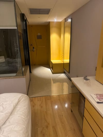 The image shows the interior of a room with a bed covered in white bedding in the lower right corner. A glass-topped surface runs along the right and bottom edges of the image, suggesting it might be a desk or table. Above it is a wooden structure with horizontal slats and a small decorative paper.

To the left is a doorway leading into a closet or storage area with light-colored wooden shelving inside. The room also has a white wall with a square vent visible.

Given the furniture, it could be a room in a hotel or an apartment. Without additional information, it's not possible to definitively place the image in Addis Ababa, Ethiopia.