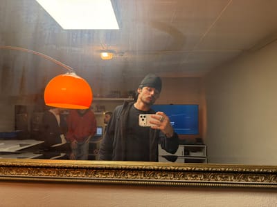The image is a mirror selfie taken indoors within a building in Oberhausen, Germany. The primary subject, a male wearing a black beanie and dark clothing, holds a white smartphone with multiple rear cameras to photograph his reflection in an ornate gold-framed mirror. He exhibits rings on his left hand and a slight pout. Reflected in the background are three additional males: one in a dark top, another in a red hoodie and blue jeans, both positioned near a large white table, and a third partially visible near an open laptop. The room features a wide-screen television displaying a blue graphical interface, situated on a white media console. Overhead lighting includes a large rectangular ceiling panel, a smaller round ceiling light, and a prominent illuminated orange spherical pendant lamp suspended by a curved metal arm. The walls are light-colored, and the ceiling shows visible paneling. The mirror's surface displays smudges and dust.