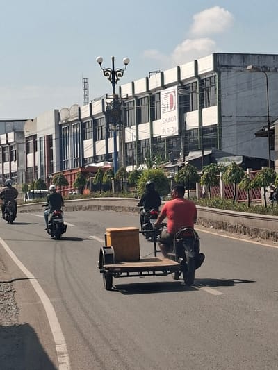 In Bireuenmeunasahcapa, Indonesia, a convoy of motorcycles travels down a paved road under a clear sky. The lead motorcycle, ridden by a person in a red shirt, is pulling a two-wheeled trailer with a wooden box. Behind them, two other motorcycles are visible, each carrying a single rider. The road is lined with a concrete barrier and decorative fencing, beyond which are trees and a multi-story building with a prominent sign reading "SHUNDA PLAFON." A cell tower is visible in the distance. The scene appears to be a typical urban street on a sunny day.
