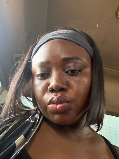 This is a close-up, overhead selfie of a young Black woman indoors, likely in a vehicle given the reflection of what appears to be a window or sunroof above and to the right of her head. She is wearing a grey headband that holds back her dark, shoulder-length hair. Her head is tilted, and she is looking directly at the camera with a neutral expression. She has a small nose ring. She is wearing a black top, and a patterned fabric, possibly a scarf or clothing, is visible draped over her shoulder. The lighting suggests it could be daytime, with natural light coming from above. The scene is otherwise nondescript, with a muted, earthy background that blends into the shadows. No text is visible in the image.
