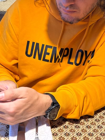 The image shows a person wearing a yellow hoodie with the word "UNEMPLOY" printed on it. The person's face is partially visible, showing a beard. They are lying on a surface with a patterned tablecloth. A watch is on the person's left wrist. Cutlery is placed on a white napkin next to the person's hand. The location is not identifiable as Ketoshi, Russia, or any other specific location.