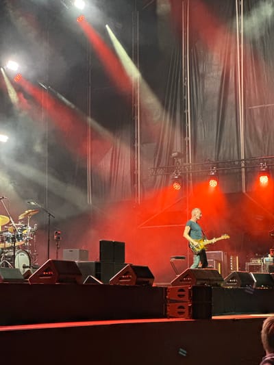 The image captures a live music performance on an outdoor stage in Chiclana de la Frontera, Spain. A male musician, likely a bassist, is the central figure, dressed in a dark t-shirt and jeans, playing a yellow electric guitar. He stands on a raised stage, illuminated by numerous red and white stage lights that cast dramatic beams through the smoky atmosphere. To the left, a drum kit is visible, with cymbals and drums catching the light. The background consists of a large black curtain and scaffolding, typical of a concert setup. Several large black speakers are positioned on the stage, both in the foreground and mid-ground. In the lower right corner, the back of a person's head, with light-colored hair, is visible, suggesting an audience member is present. The overall scene is one of a dynamic and energetic live musical event.