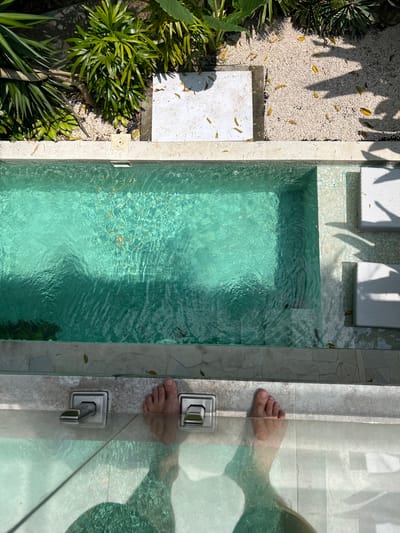 A top-down shot shows a small, rectangular pool filled with clear, turquoise-tinted water. The pool is surrounded by beige stone tiles. Adjacent to the pool, on one side, is a gravel area with scattered leaves and a patch of green foliage. Two white seating structures are positioned near the pool on the same side as the gravel. On the opposite side of the pool, a glass barrier is visible, partially obscuring a person's feet, standing at the pool's edge, indicating someone entering or exiting. The scene appears to be set outdoors, possibly in a private garden or resort. Based on the metadata, this image was taken in Buduk, Indonesia.
