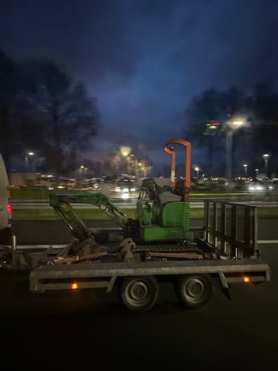 The image captures a nighttime scene in Hoofddorp, Netherlands, depicting a green compact excavator secured on a metallic gray dual-axle flatbed trailer. The excavator features an orange roll-over protective structure (ROPS) and "bam" branding on its green boom. The trailer displays two illuminated orange side marker lights and a dark, lattice-patterned metal gate or ramp at its rear. A white van, with its red left taillight visible, is partially observed on the far left, consistent with towing the trailer. The vehicles are positioned on a paved multi-lane road. The background reveals active traffic with numerous vehicle headlights and taillights appearing as blurred streaks of light, indicating motion. Tall streetlights emit yellow-orange light, and a traffic signal displaying red and green lights is visible in the upper right. Dark, blurred trees and atmospheric smoke or fog are present in the distant background, under a dark blue-black sky.