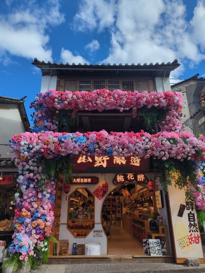 This media file captures a vibrant scene in Dali City, China, likely during the daytime under a bright, cloudy sky. The primary focus is a building entrance elaborately decorated with abundant floral arrangements in shades of pink, red, blue, and purple. These flowers form archways and cascades, creating a festive and welcoming atmosphere.

The building itself appears to be a traditional structure with tiled roofs and wooden window frames. The storefront visible behind the floral displays is a shop selling alcoholic beverages, as indicated by Chinese text. One prominent sign reads "大理老酒坊" (Dali Old Wine House) and another "桃花醉" (Peach Blossom Drunk), suggesting a specialty wine shop. Other visible text includes "山野酿造" (Wilderness Brewing), "手作" (Handmade), and information about hiring a salesperson.

The overall impression is one of a lively marketplace or a tourist-oriented establishment in Dali, known for its cultural heritage and scenic beauty. The bright flowers and clear sky evoke a sense of celebration or a pleasant day for visiting the local shops. There are no people clearly visible in the foreground, but reflections in the shop window hint at activity inside.