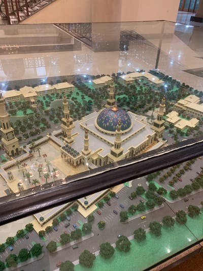 This image captures an overhead view of a meticulously crafted architectural model, likely depicting a large mosque complex. The model is displayed under a clear glass case, which reflects the indoor surroundings, including ceiling lights, structural elements, and a staircase.

The central and most prominent subject of the model is a grand mosque, featuring a large, intricately decorated dome in blue and gold patterns, flanked by multiple tall minarets. Surrounding the main mosque structure are numerous smaller, beige-colored buildings, integrated into the complex. The model also includes extensive landscaping, with miniature trees, bushes, and pathways, giving a sense of a well-planned urban environment. Miniature cars are parked in designated lots and line the roads, some with tiny streetlights. A small piece of text visible on one of the miniature roads reads "JL. ANDI."

The scene is a static display, showcasing the detailed design and layout of the complex. Given the location context of Samarinda, Indonesia, this model is highly likely to represent the Samarinda Islamic Center Mosque, known for its impressive scale and distinct architecture. The presentation suggests it is a permanent exhibit within an indoor setting, such as a museum, visitor center, or the complex itself, designed to offer an overview of the entire facility. No people are visible within the model or in the reflections, maintaining the focus on the architectural work.