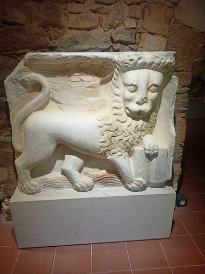A stone relief of a winged lion is displayed on a light-colored rectangular pedestal against a rough stone wall in a museum in Chalkida, Greece. The lion, carved in white stone, is depicted in profile, facing left, with its body elongated and muscular. It has a mane of wavy curls, large expressive eyes, and a long, curled tail. Its front paws rest on an unseen object, and it appears to be standing on a wavy, textured surface. The background wall is composed of irregularly shaped, rough-hewn stones in varying shades of brown and gray. The floor is tiled with reddish-brown square tiles. A small black electrical box is visible on the floor to the right of the pedestal. A label is affixed to the front of the pedestal, though its text is not legible.