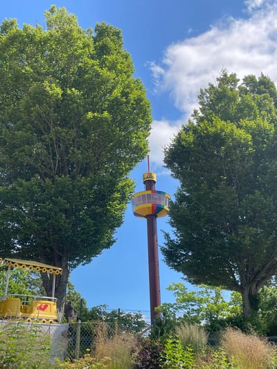 This is a full-frame, eye-level, outdoor, vertical shot of a colorful observation tower in the city of Billund, Denmark, in front of a bright blue sky with scattered white clouds. The tower, branded with the LEGO logo and the word "OLAND," features a circular observation deck with a rainbow-colored band around its base. Two large, mature green trees flank the tower, framing it in the center of the composition. In the foreground, to the left, a yellow amusement park ride, resembling a train car with a decorative canopy, is visible behind a chain-link fence. The area is lush with greenery, including tall grasses and shrubs, and a fence runs across the middle ground. The scene depicts a sunny day in an amusement park setting.