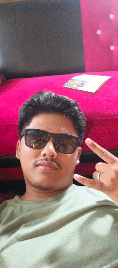 A young man wearing sunglasses and a light green t-shirt is pictured from the chest up, posing for a selfie. He has dark hair and a mustache. His right hand is raised, forming a peace sign with his index and middle fingers, while his left hand is partially visible. A silver ring is on his left ring finger. He is positioned in front of a vibrant pink surface, which appears to be a sofa. To the right of the man, a small, open booklet with visible illustrations is placed on the pink surface. The background consists of a dark, textured surface above the pink one. The lighting suggests an indoor setting. The overall impression is a casual, personal photograph taken in a home environment.