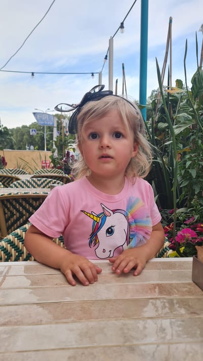A young, fair-skinned child with blonde hair and a black headband is standing next to a tiled table. She is wearing a pink t-shirt with a unicorn graphic. The background includes green foliage, flowers, and part of a blue structure. Street signs are visible in the background. The location is Kiryat Gat, Israel.