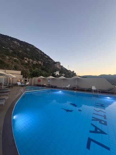 The image depicts an outdoor swimming pool at the Mistral Hotel in Agios Nikolaos Municipal Unit, Greece. The pool is filled with bright blue water and features a tiled bottom. The word "MISTRAL" is visible in large, dark letters at the bottom of the pool, and two dark silhouettes of dolphins are also present. The pool area is surrounded by lounge chairs and umbrellas. In the background, a steep, rocky hillside covered with vegetation rises behind the hotel buildings. The sky is clear and a pale blue, suggesting it is either early morning or late evening. The scene appears peaceful and serene.