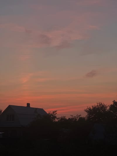 A photograph captured at dusk in Krivandino, Russia, depicts a serene rural landscape. The sky is awash with the soft hues of sunset, transitioning from a warm orange near the horizon to a pale pink and lavender at the zenith. Wispy clouds are scattered across the sky, adding texture and depth to the celestial canvas.
In the foreground, the silhouette of a two-story house stands prominently. The house is dark against the illuminated sky, with a steeply pitched roof and a single window visible on the upper floor. A chimney rises from the peak of the roof, also silhouetted. The house appears to be surrounded by dense foliage, forming a dark mass that obscures the lower portion of the structure and extends to the sides.
To the right of the house, the dark shapes of trees create a dense treeline. The overall impression is one of quiet solitude, with the fading light casting long shadows and emphasizing the peaceful ambiance of the countryside. The image is a still life, capturing a moment of natural beauty and the simple charm of a rural dwelling at the close of day.