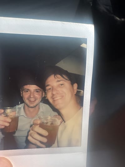Two Caucasian males are pictured in a close-up, indoor, candid photograph, likely taken with a flash. The man on the left, wearing a light blue collared shirt and a dark cap, is holding a clear plastic cup filled with a brown beverage. He is smiling broadly and looking towards the camera. The man on the right, wearing a light-colored t-shirt, is also smiling and looking towards the camera, holding a similar clear plastic cup with a brown beverage. The background is dark and indistinct, with some bright light sources visible, suggesting artificial lighting. The photograph is framed by a white border typical of instant film. The scene is devoid of any recognizable landmarks or environmental clues that would allow for a definitive identification of the location within Hallandale Beach, United States of America.