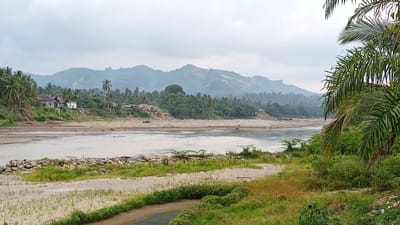 This image depicts a hazy, overcast day in Beunyot, Indonesia, with a wide river dominating the foreground. The river's banks are lined with sand and sparse vegetation, with a line of large rocks forming a barrier on the near bank. In the midground, a dense jungle of palm trees and other foliage covers the landscape, interspersed with a few buildings, including houses with corrugated metal roofs. A dirt road with patches of grass winds through the sandy area in the foreground. In the background, mist-shrouded mountains rise, their slopes covered in vegetation. The overall impression is one of a rural, tropical environment.