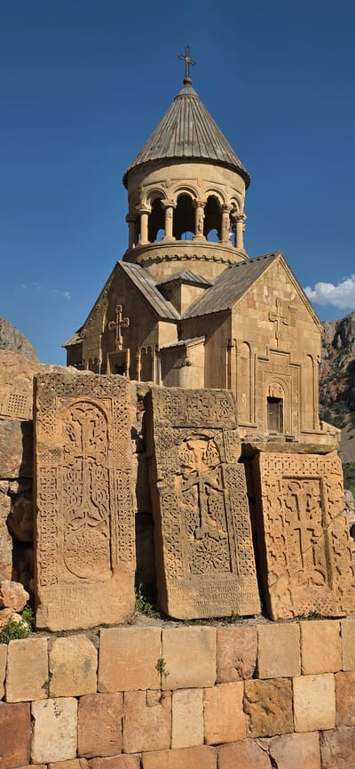 The image displays the Noravank Monastery complex located in Amaghu, Armenia. In the foreground, three large, intricately carved stone slabs, known as khachkars, stand upright. These khachkars are ancient Armenian cross-stones, each adorned with elaborate knotwork and Christian symbols. Behind the khachkars, the monastery building is visible, characterized by its weathered sandstone construction. The main structure features a conical dome topped with a cross, supported by an octagonal drum with arched openings. The facade of the building is embellished with crosses and decorative carvings. The surrounding environment includes rocky hills and a clear blue sky.