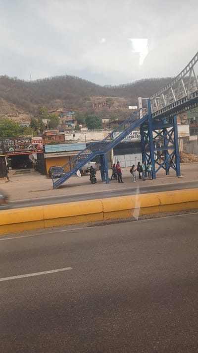 The image depicts an outdoor scene in Puerto La Cruz, Venezuela, featuring a blue pedestrian bridge crossing over a roadway. Several people are standing near the bridge, and motorcycles are parked nearby. The presence of a building labeled "Racing Auto Accesorios" suggests a commercial area. The background includes a hillside with vegetation and scattered buildings. The roadway has a yellow curb.