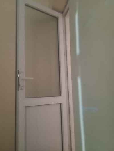 The image shows a white door with a glass panel at the top and a solid panel at the bottom. The door has a silver handle. The door is set within a white frame, and it appears to be an interior door. The wall to the left of the door is a neutral beige color, and the wall to the right is a light green color. There are two vertical light reflections on the green wall, suggesting a light source is present. The image quality is poor, with low resolution and an out-of-focus appearance. No people or specific actions are visible in the image. The scene appears to be an indoor setting, possibly a hallway or room, with a door leading to another area. No distinctive features that would allow for precise identification of the location within Kyzyltepa, Uzbekistan are present.