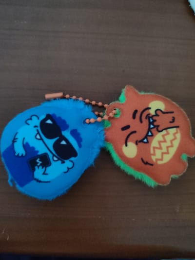 Two fuzzy monster-shaped keychains are laid out on a brown wooden surface. The keychain on the left is primarily blue and features a cartoon monster wearing black sunglasses and holding a small rectangular object, possibly a phone. The monster has white fangs and round yellow cheeks. The keychain on the right is orange and has a fuzzy green trim. It depicts a cartoon monster with its eyes closed, mouth open with white fangs, and one hand covering its face. It also has yellow circles for cheeks and a yellow zig-zag pattern on its body. A beaded orange string connects the two keychains. The lighting suggests it is daytime, and the setting appears to be indoors, possibly on a table. No visible text is present.