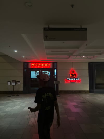 The photograph depicts the entrance to the "Volcano Shabu Shabu & Grill" restaurant in Putrajaya, Malaysia. The signage for the restaurant is illuminated in red neon. A sign indicates the location is UG (Underground) level, near the "Centre Court". A person holding a phone is partially visible in the foreground. The environment appears to be a mall or commercial center.