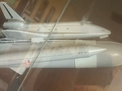 The image displays a detailed model of the Space Shuttle system, captured from a slightly elevated side view. The main subjects are the orbiter (space shuttle), a segment of the large external tank, and one of the solid rocket boosters.

The model is predominantly a light grey or silver color, featuring intricate panel lines and structural details. A prominent detail is a small American flag decal affixed to the side of the solid rocket booster. The nose cone of the orbiter is visible, showing small circular indentations that could represent windows or sensors. Towards the rear, the orbiter's engine nozzles and a portion of its vertical stabilizer are discernible.

The model appears to be housed within a clear display case or behind a protective barrier, as indicated by reflections and diagonal lines across the image, suggesting glass or plastic. The background is blurred and dark, consisting of brown and orange tones, consistent with an indoor setting, possibly a museum, private collection, or retail display. No explicit activity is taking place, as it is a static model on exhibition.

While the location context of Yana, Bulgaria, is provided, the image itself does not contain any visual cues specific to this geographical location; it solely depicts the space shuttle model in an indeterminate indoor setting.